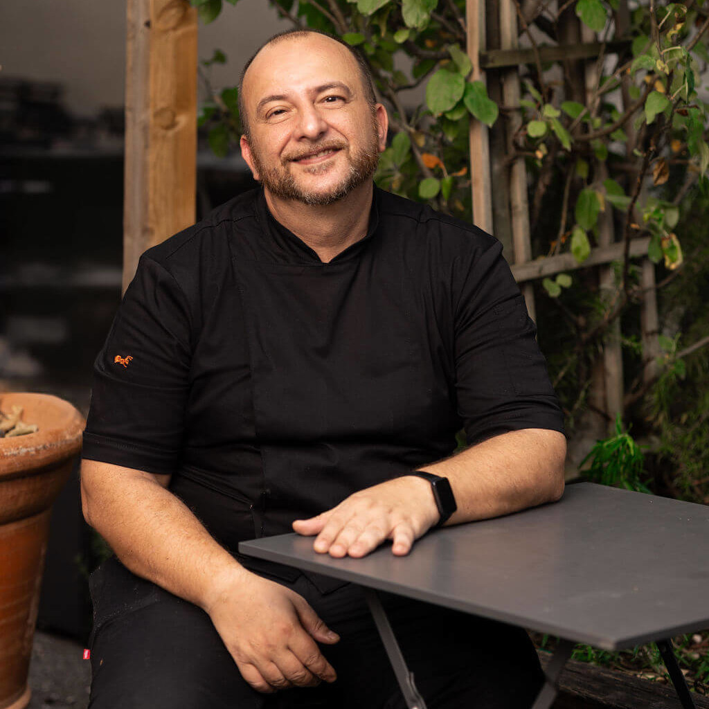 Portrait d'un chef souriant, vêtu d'une tenue noire, assis à une table extérieure entourée de plantes en arrière-plan.