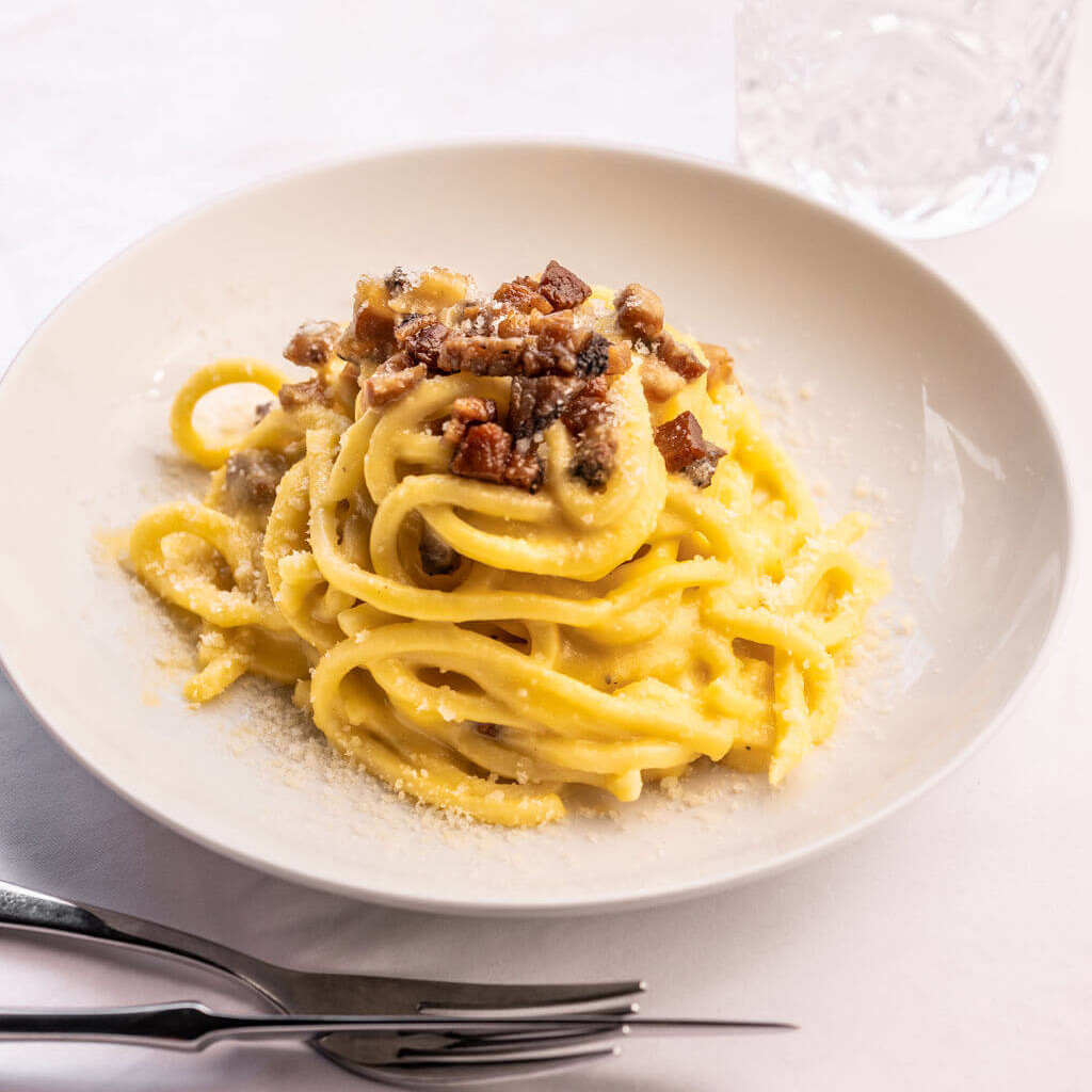 Assiette de carbonara crémeuse avec lardons croustillants et parmesan, accompagnée de couverts élégants sur une table blanche.