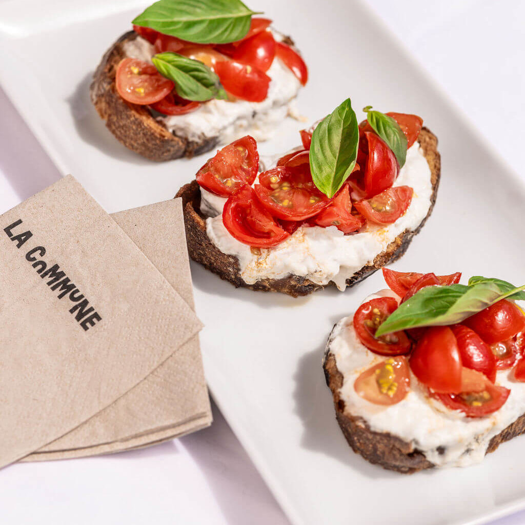 Tartines garnies de straciatella , tomates cerises et basilic sur pain grillé, servies à La Commune avec serviette en papier marquée.