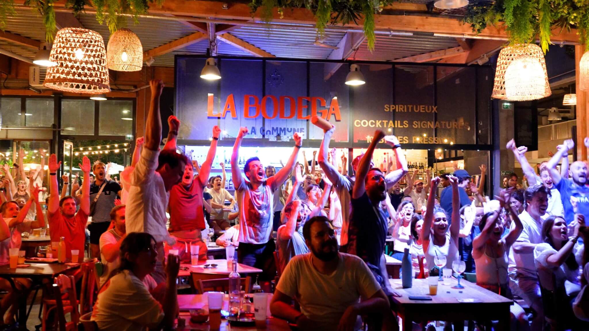 Une foule en liesse dans un bar de La Bodega, les bras levés, célébrant un moment de victoire.