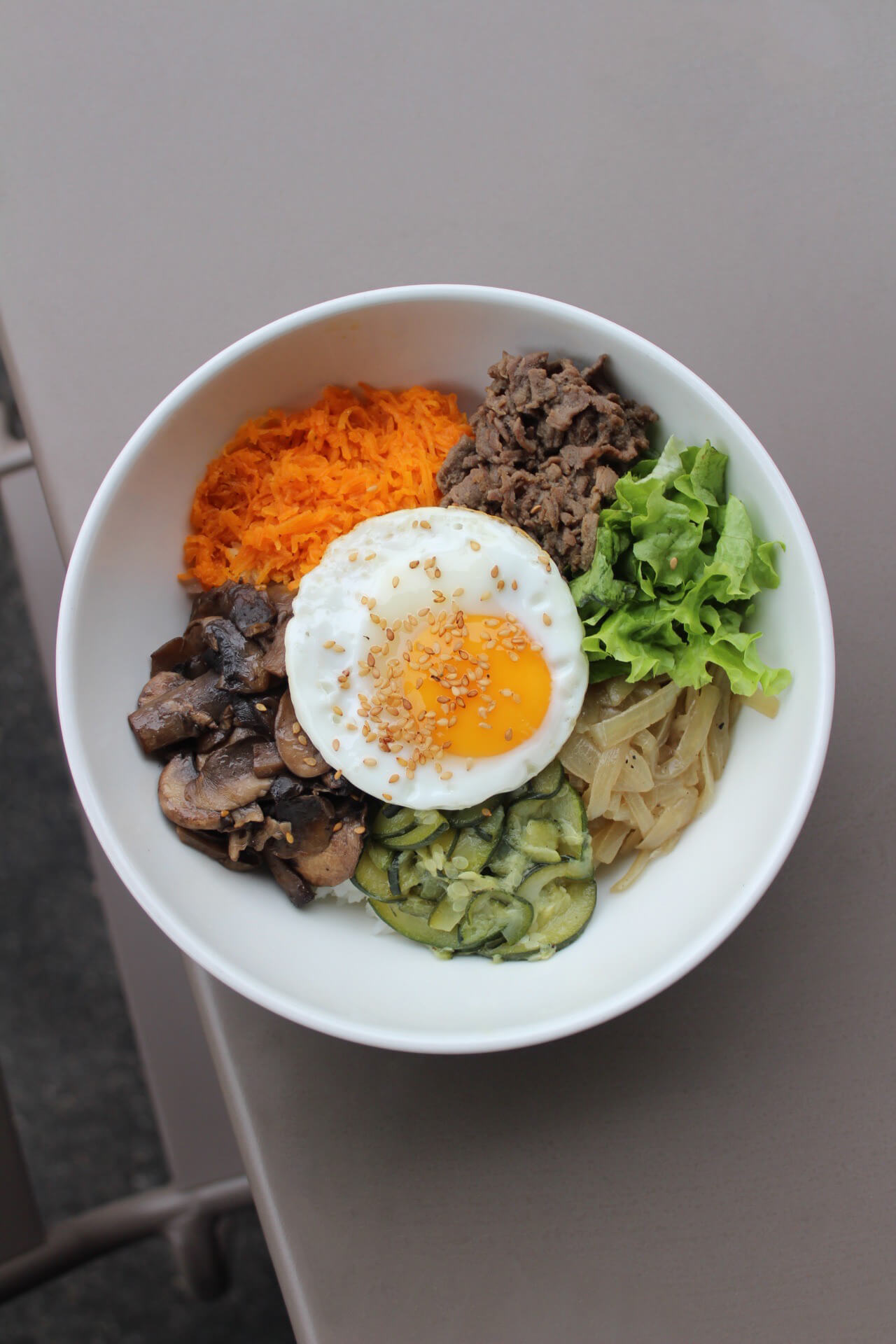 Bol de Bibimbap coréen avec œuf au plat, carottes râpées, champignons, bœuf mariné, salade verte et riz, garni de graines de sésame.