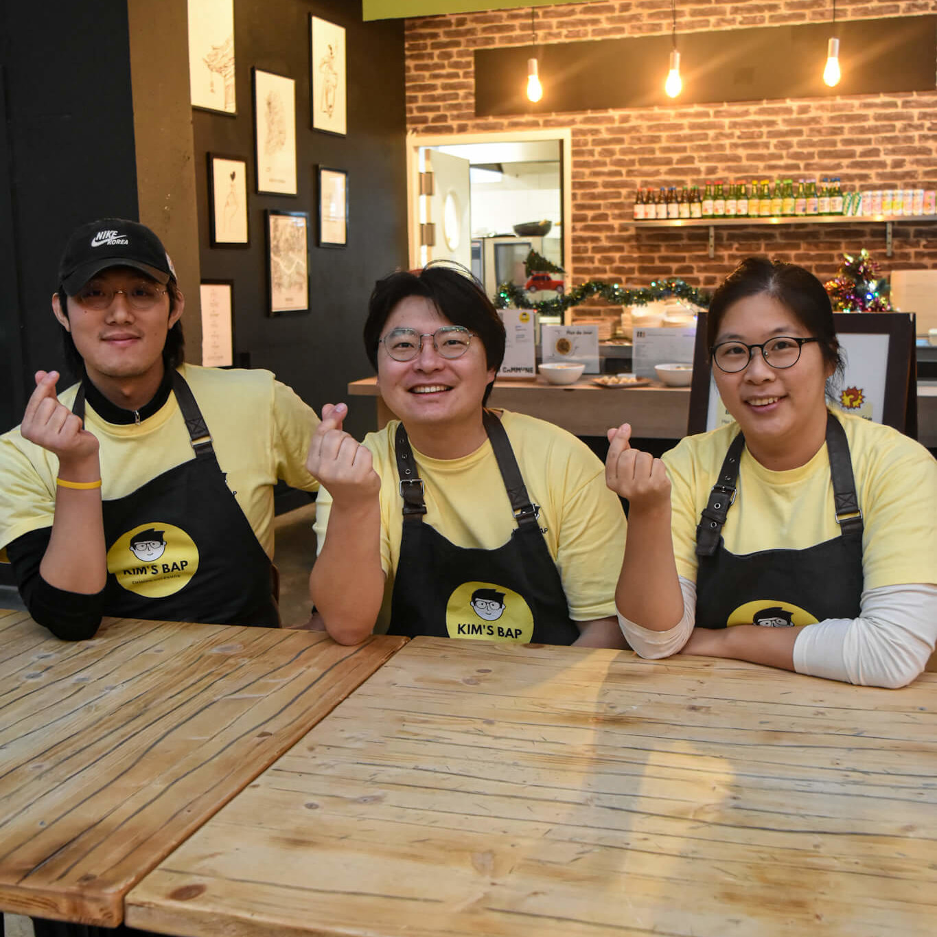 L'équipe de Kim's Bap, restaurant de cuisine coréenne, posant souriante devant leur comptoir, vêtue de t-shirts jaunes et tabliers noirs avec leur logo.