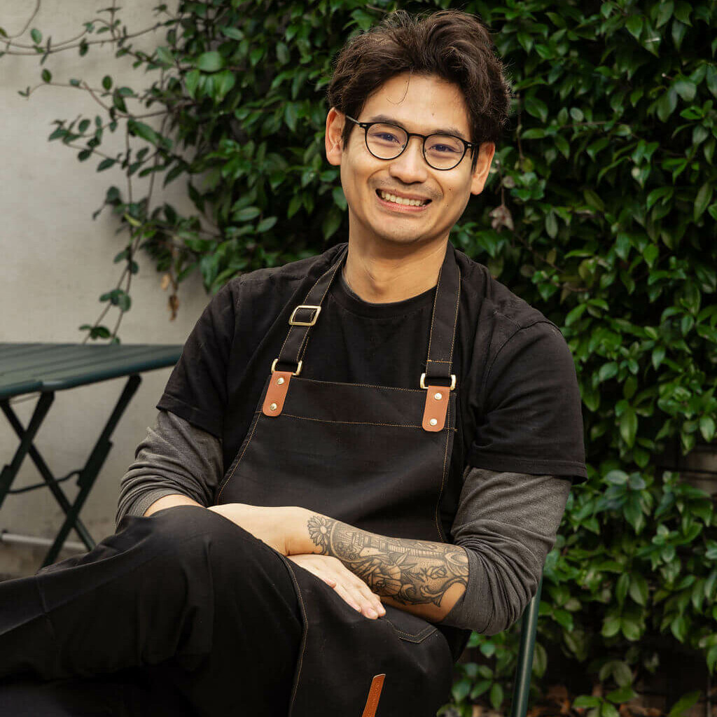Portrait d'un chef souriant assis dans un jardin, portant un tablier noir avec des lanières en cuir et des lunettes, entouré de verdure.