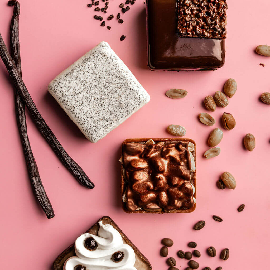 Quatre pâtisseries gourmandes présentées sur un fond rose, accompagnées de gousses de vanille, éclats de cacao, graines et grains de café.