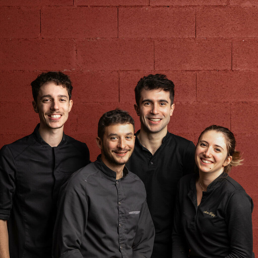Portrait d'une équipe de pâtissiers souriants posant devant un mur rouge, vêtus de tenues professionnelles noires.