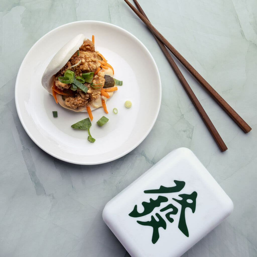 Bao garni de poulet frit, légumes croquants et ciboulette, présenté sur une assiette blanche avec des baguettes sur un fond marbré