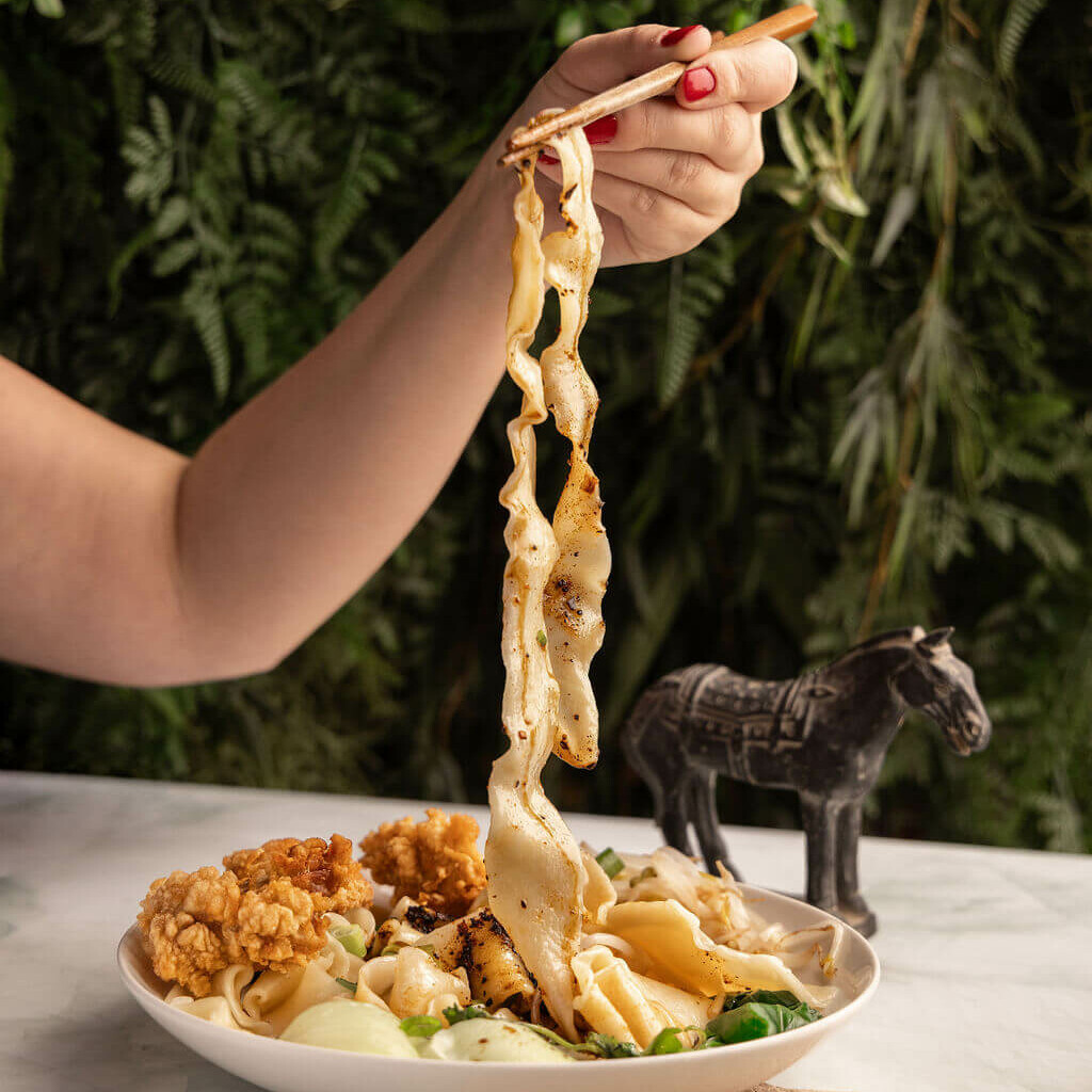Nouilles Biang Biang soulevées avec des baguettes, mettant en valeur leur texture, accompagnées de poulet frit et d'une décoration de cheval, sur fond végétal.