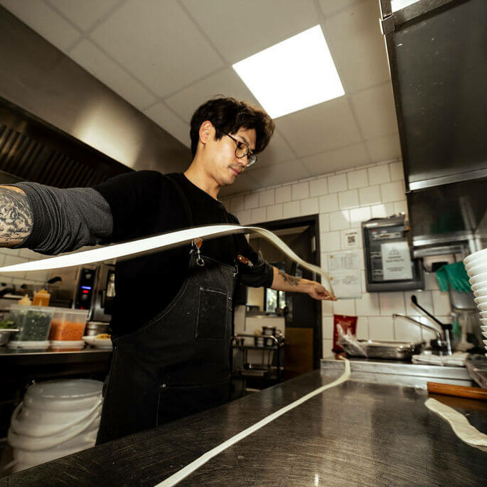 Chef étirant une longue pâte à nouilles dans une cuisine professionnelle moderne, entouré d'équipements et de bols empilés.