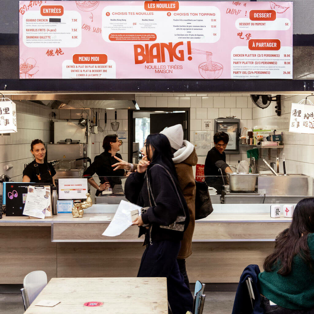 Stand de nouilles Biang! avec le menu affiché en détail, une équipe en cuisine et des clients dans un food court animé.
