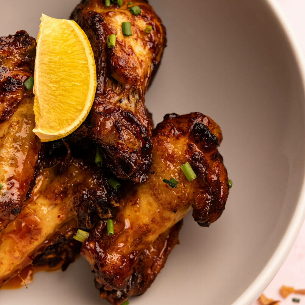 Ailes de poulet laquées d’une sauce caramélisée et garnies de ciboulette fraîche, servies avec un quartier d’orange, dans une assiette blanche épurée.
