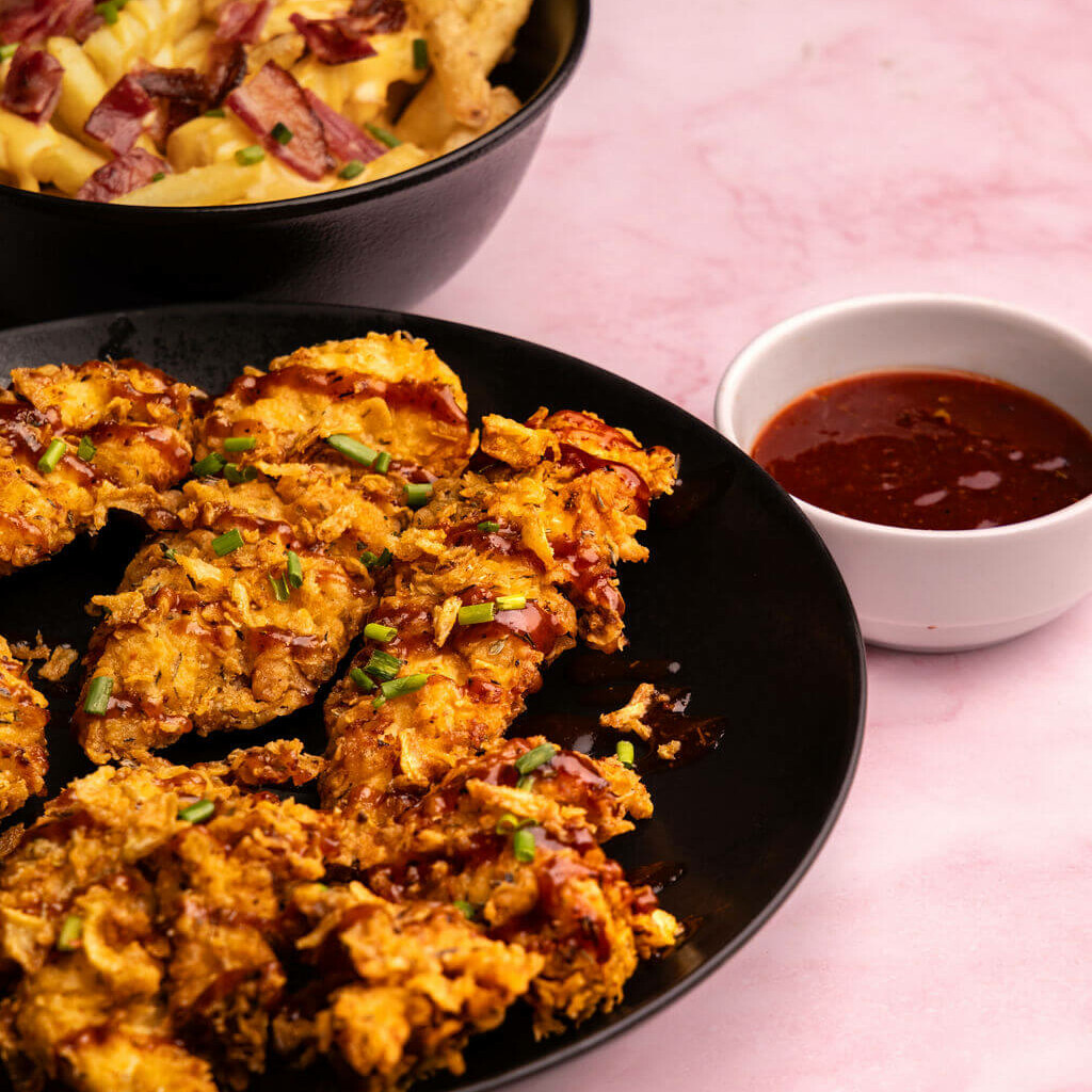 Assiette de tenders de poulet frit avec une sauce barbecue épicée, accompagnée de frites garnies de cheddar fondu et bacon croustillant, sur fond rose.