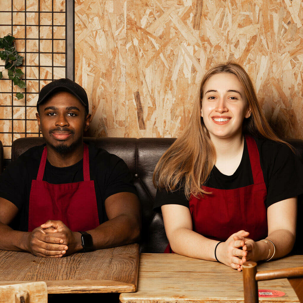 Les fondateurs de Crousty Chicken, souriants, en tablier rouge, assis dans un décor en bois chaleureux, incarnant l'esprit convivial de leur enseigne.