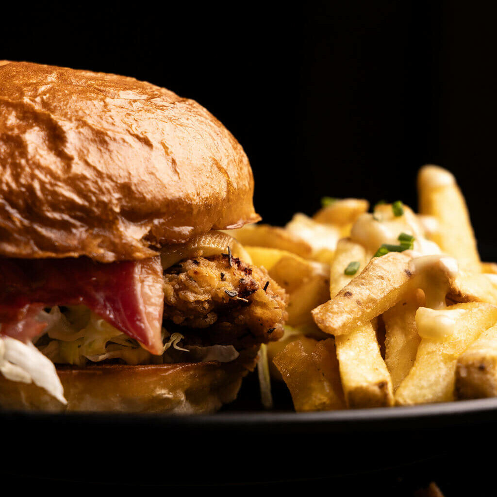 n burger au poulet croustillant avec fromage fondu et pickles, accompagné de frites généreusement nappées de sauce fromage, sur une assiette noire élégante.