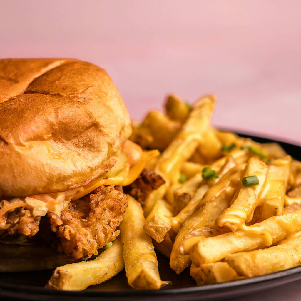 Gros plan sur un burger au poulet frit, garni de salade et de sauce maison, avec des frites croustillantes, servi sur une assiette noire sur fond sombre.